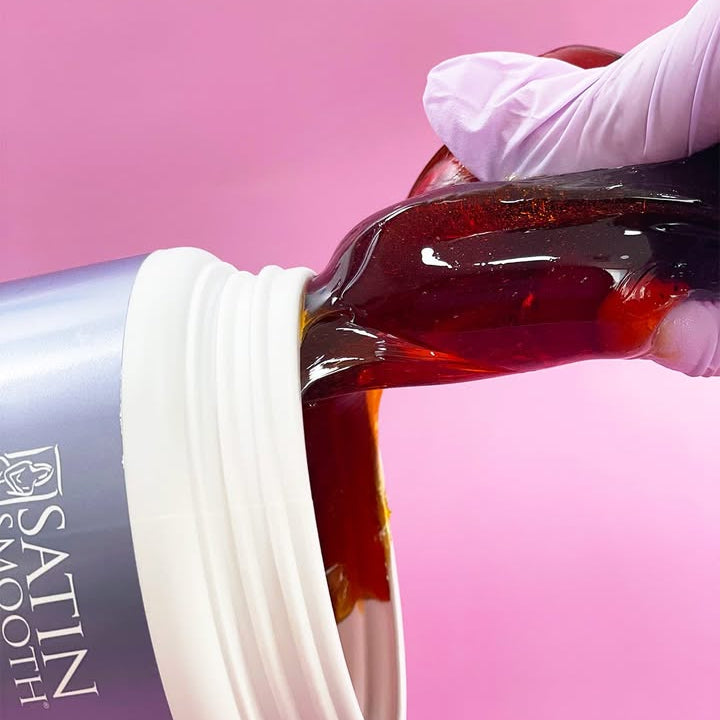 Hand pouring a bottle 'Satin Smooth' onto a white surface with a pink background.